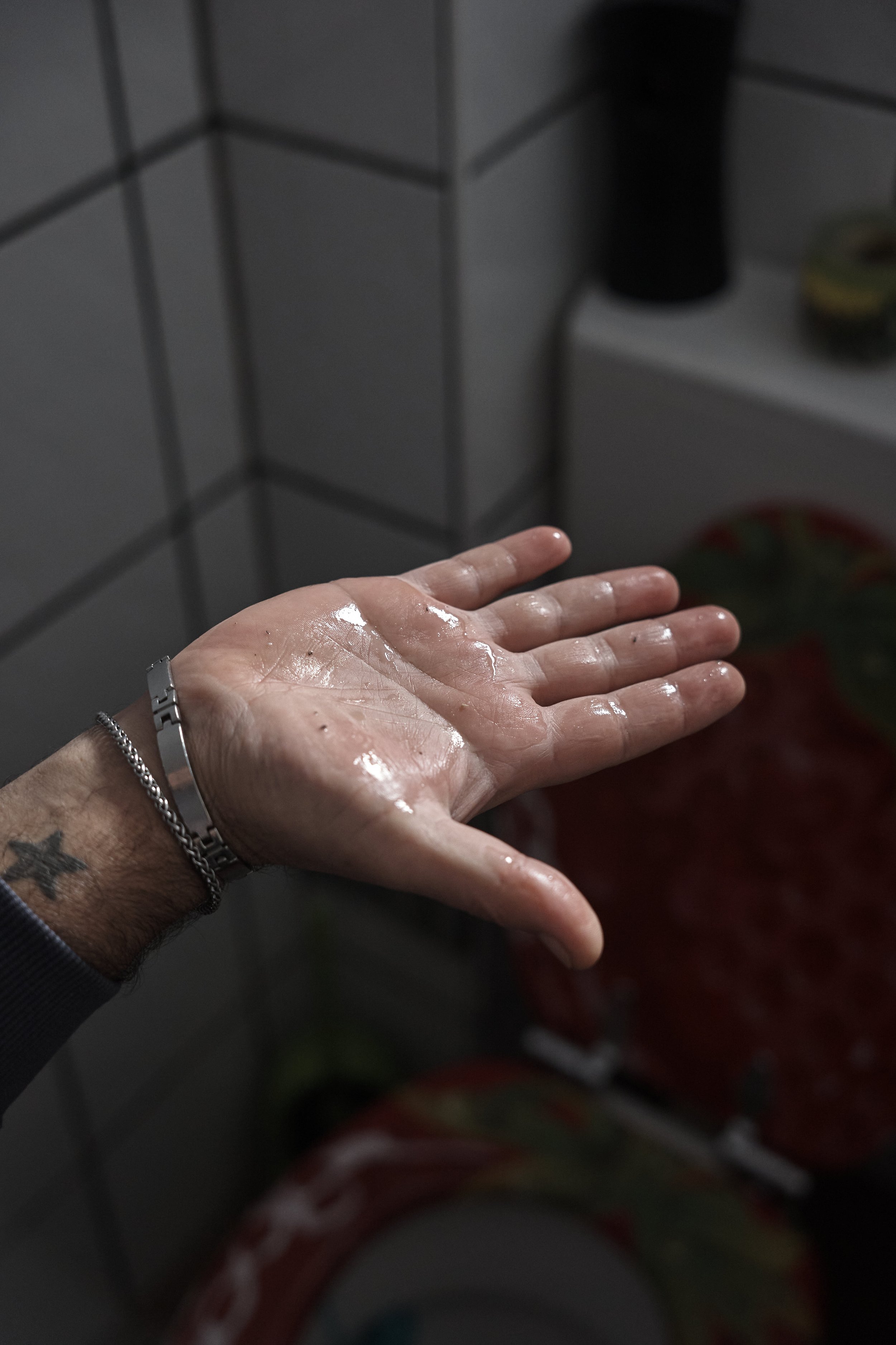 Eine tätowierte Hand mit Armband, mit Creme eingerieben, über einem gekachelten Waschbecken.