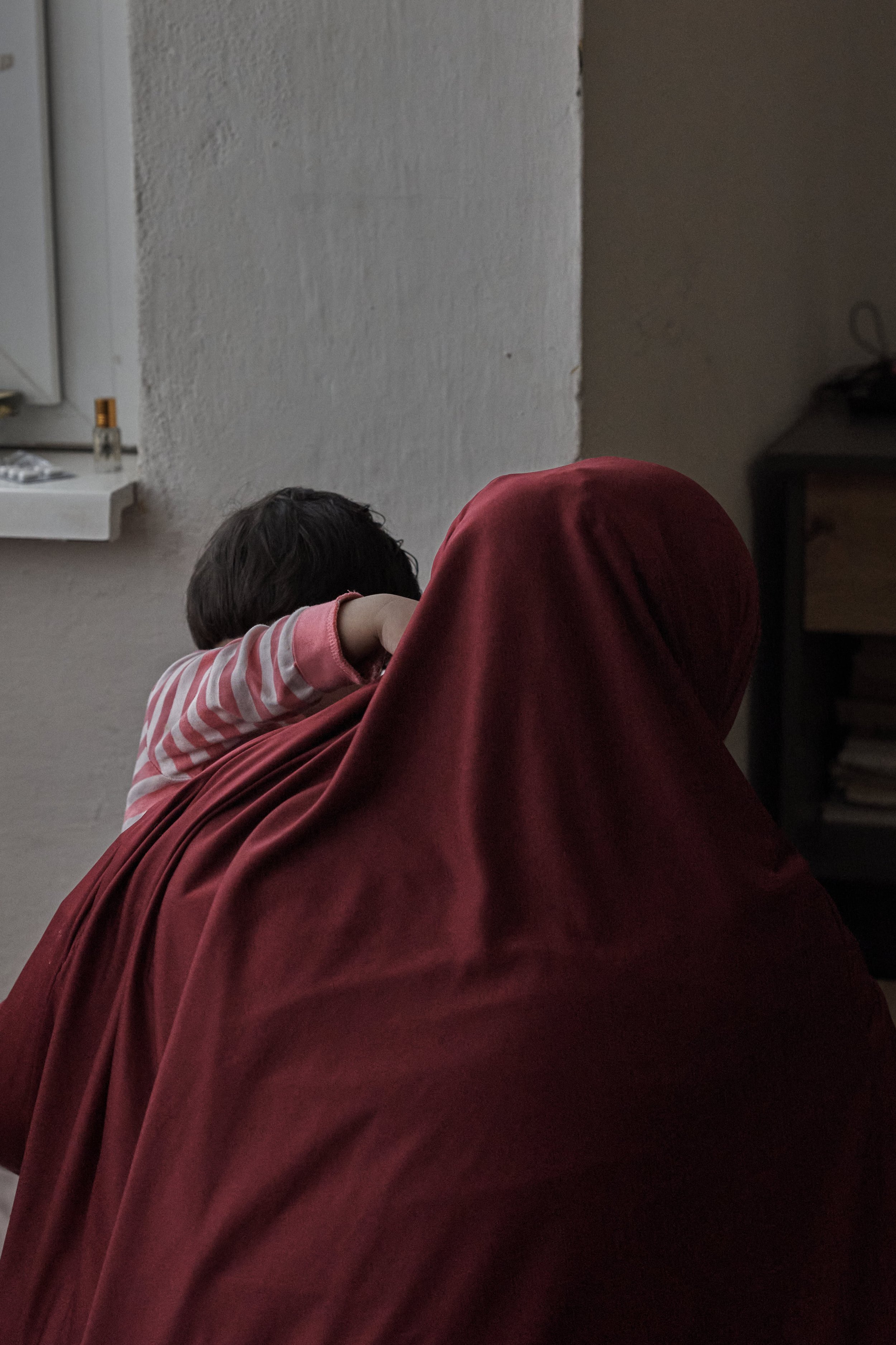 Eine Person mit rotem Kopftuch hält ein Kind im Arm, beide blicken aus dem Fenster.
